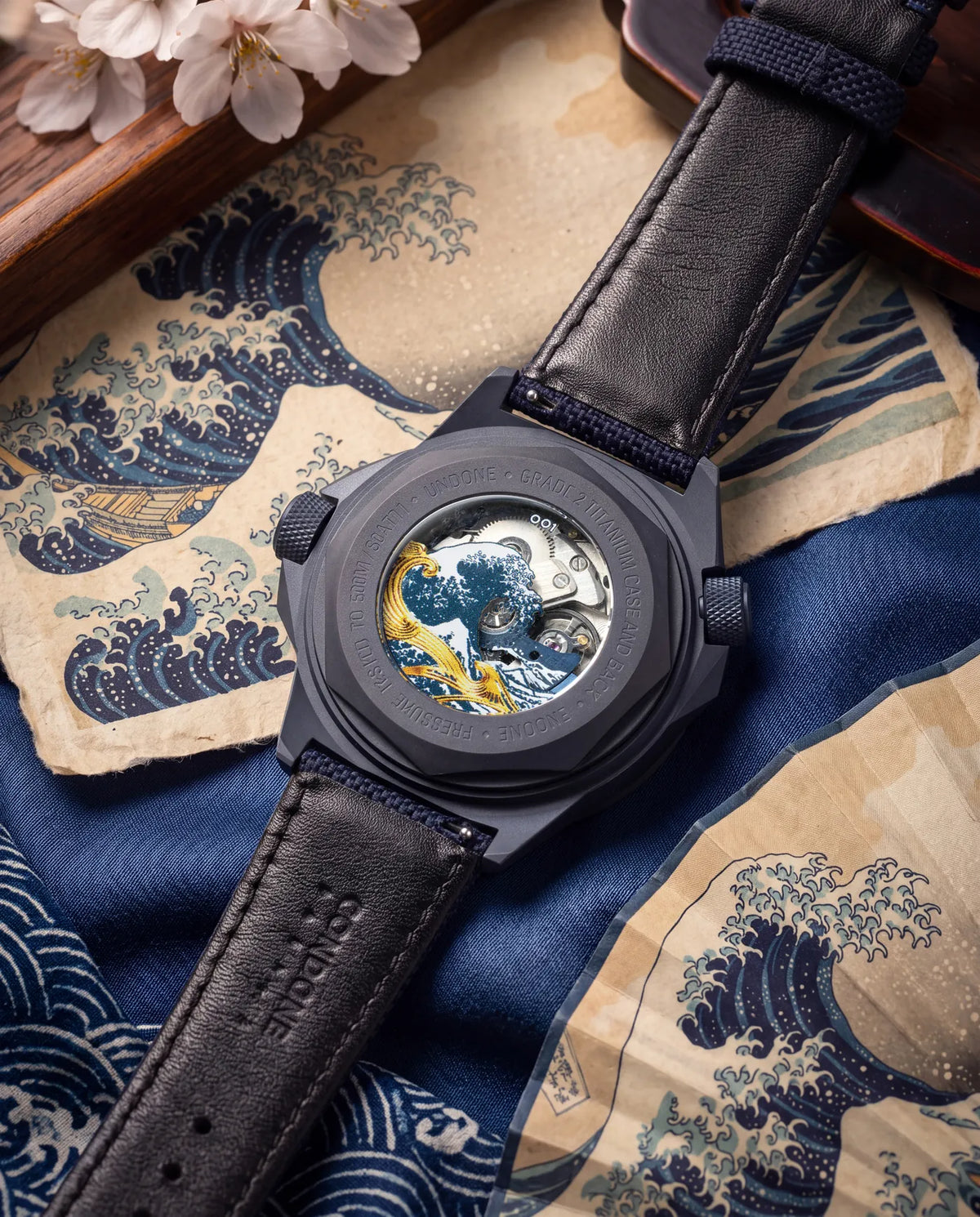 The Hokusai II caseback on its strap, laid over Great Wave prints beside cherry blossom