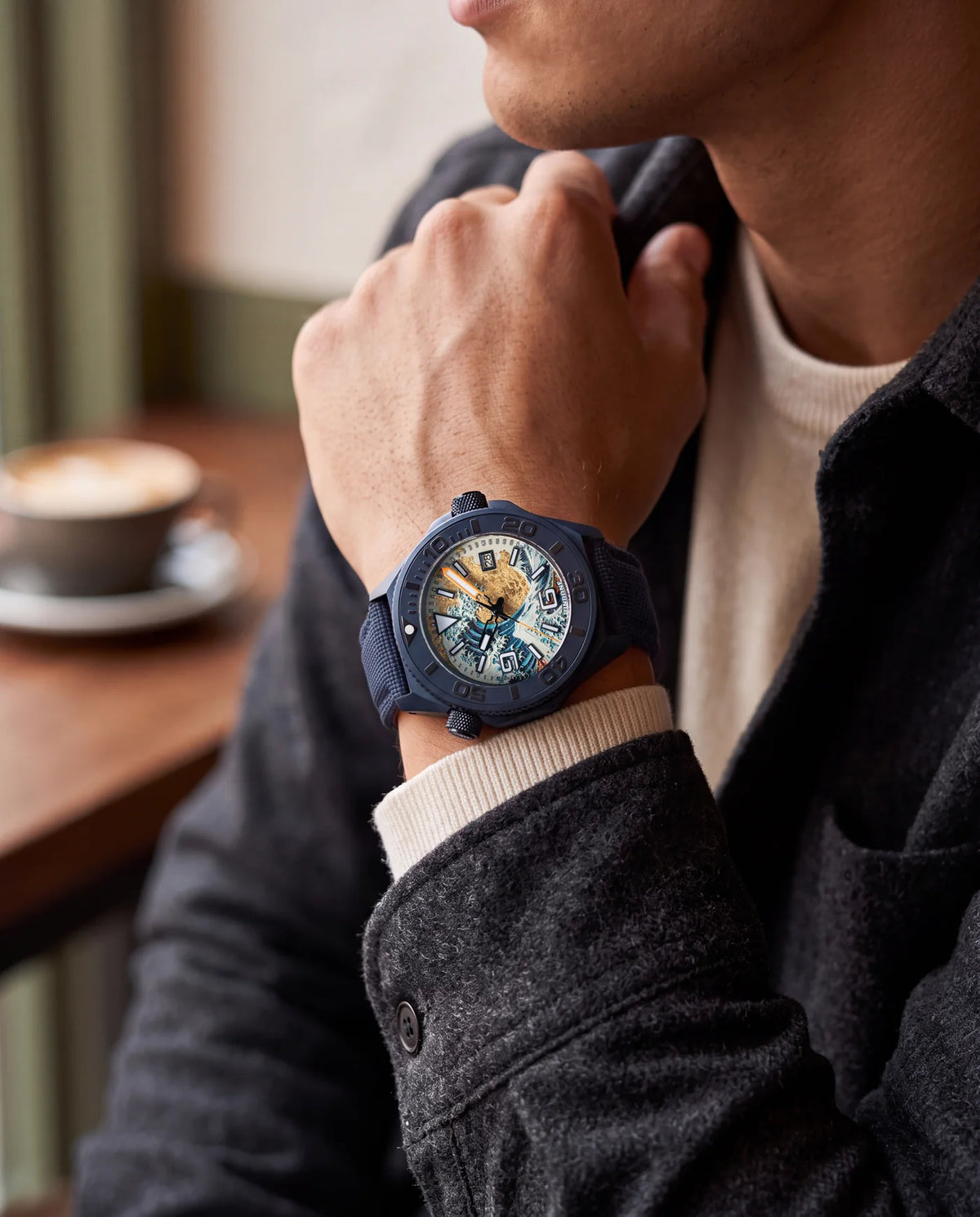 Hokusai II worn on the navy Cordura strap, a hand raised to the collar of a dark wool jacket, a coffee cup on the table behind