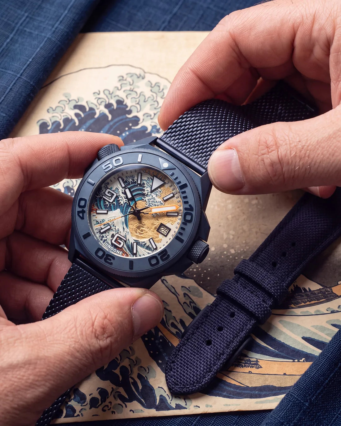 Hands fitting Hokusai II on its blue Milanese mesh, the navy Cordura strap laid beside it on a Great Wave print